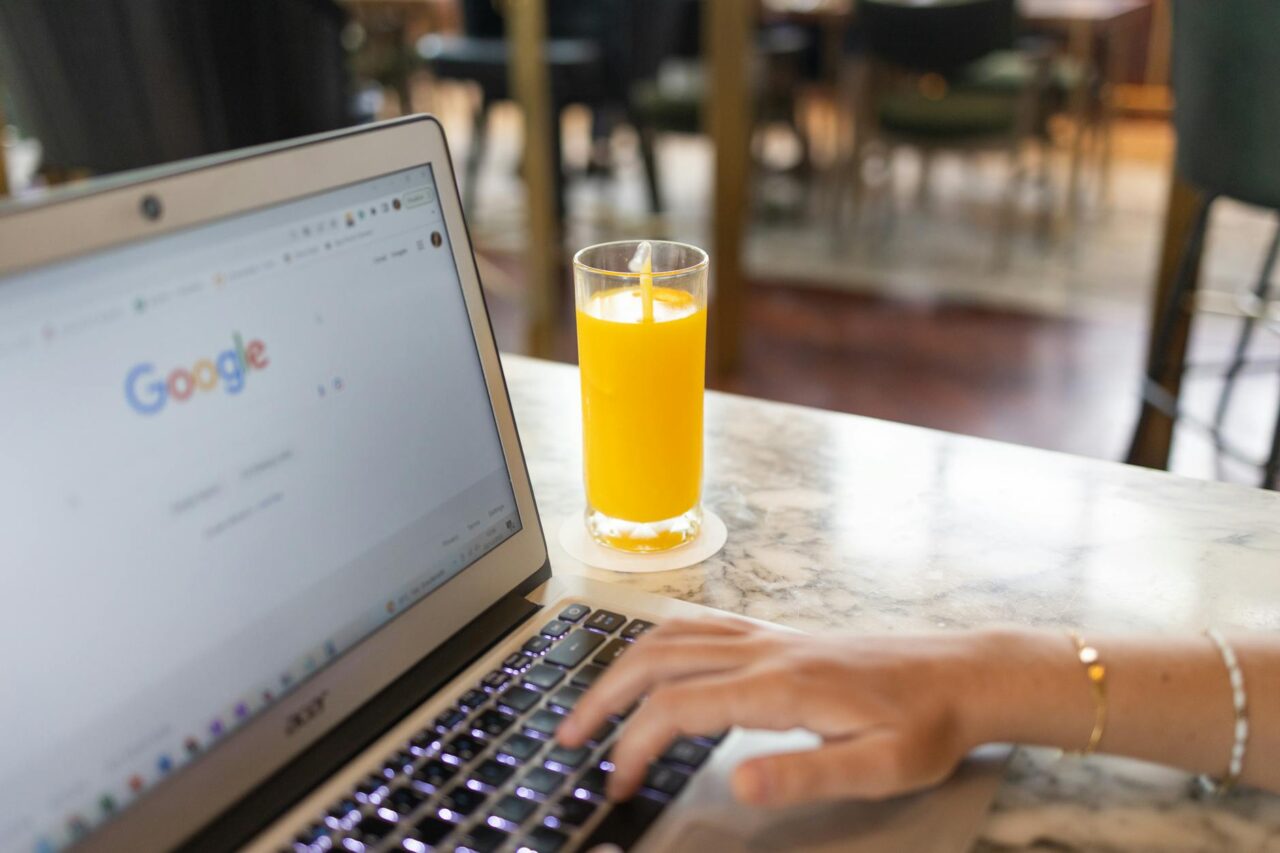 seo-boost-2T Mână care tastează pe un laptop cu pagina de căutare Google deschisă, lângă un pahar de suc de portocale